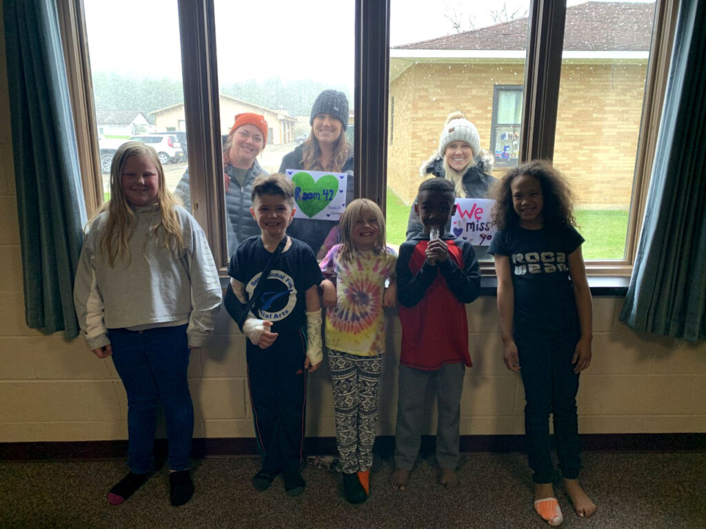 elementary students pose in front of a window with teachers in the background